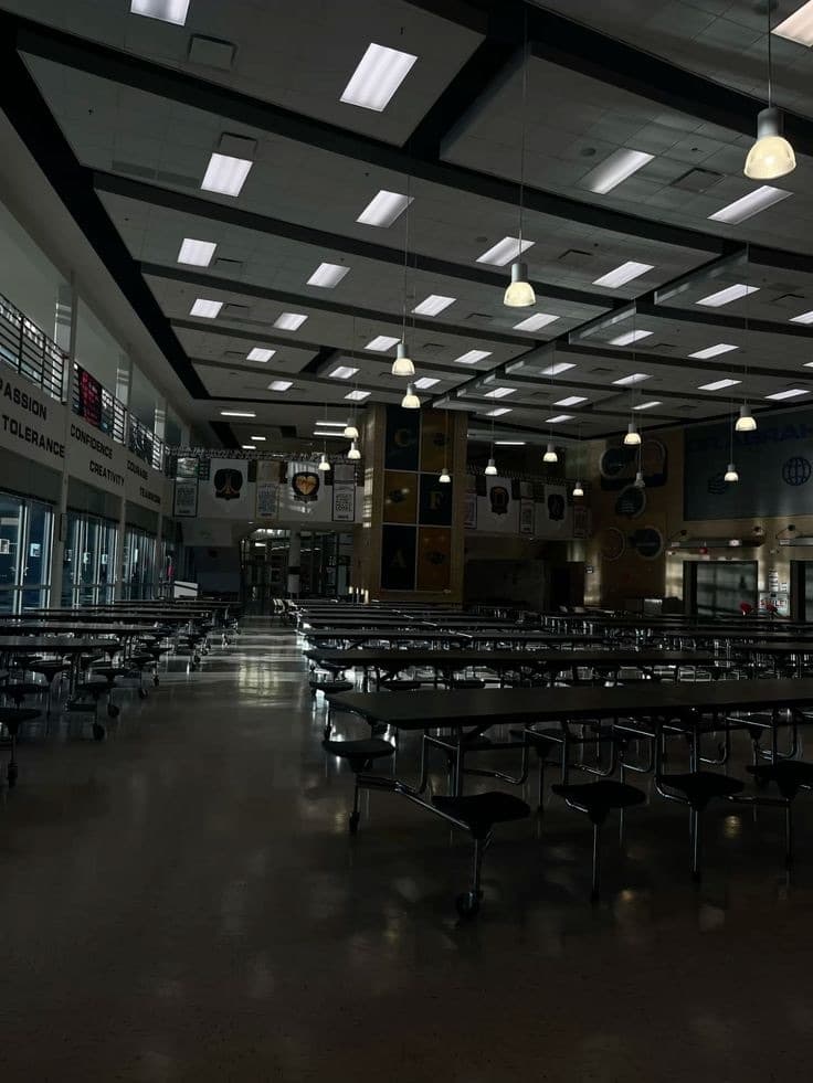 Une grande salle déserte aux longues tables alignées, éclairée par des plafonniers dans la pénombre.