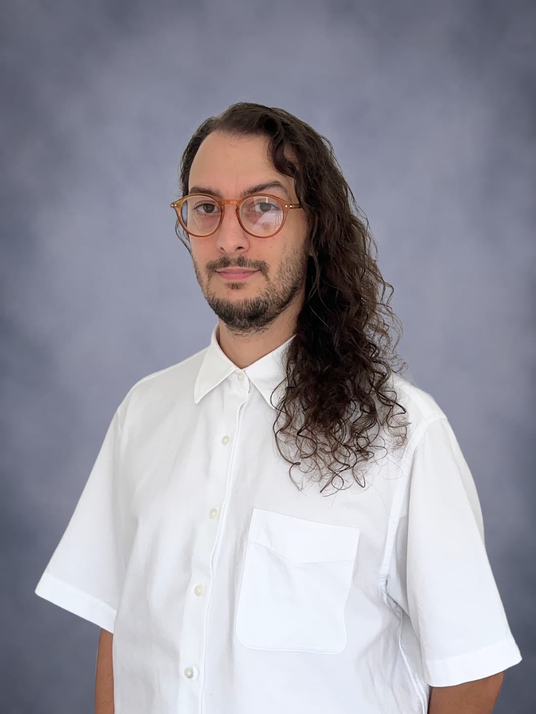 Luc Di Giorgio, cheveux bruns bouclés longs, lunettes rondes ambrées et barbe courte, en chemise blanche, devant un fond gris.